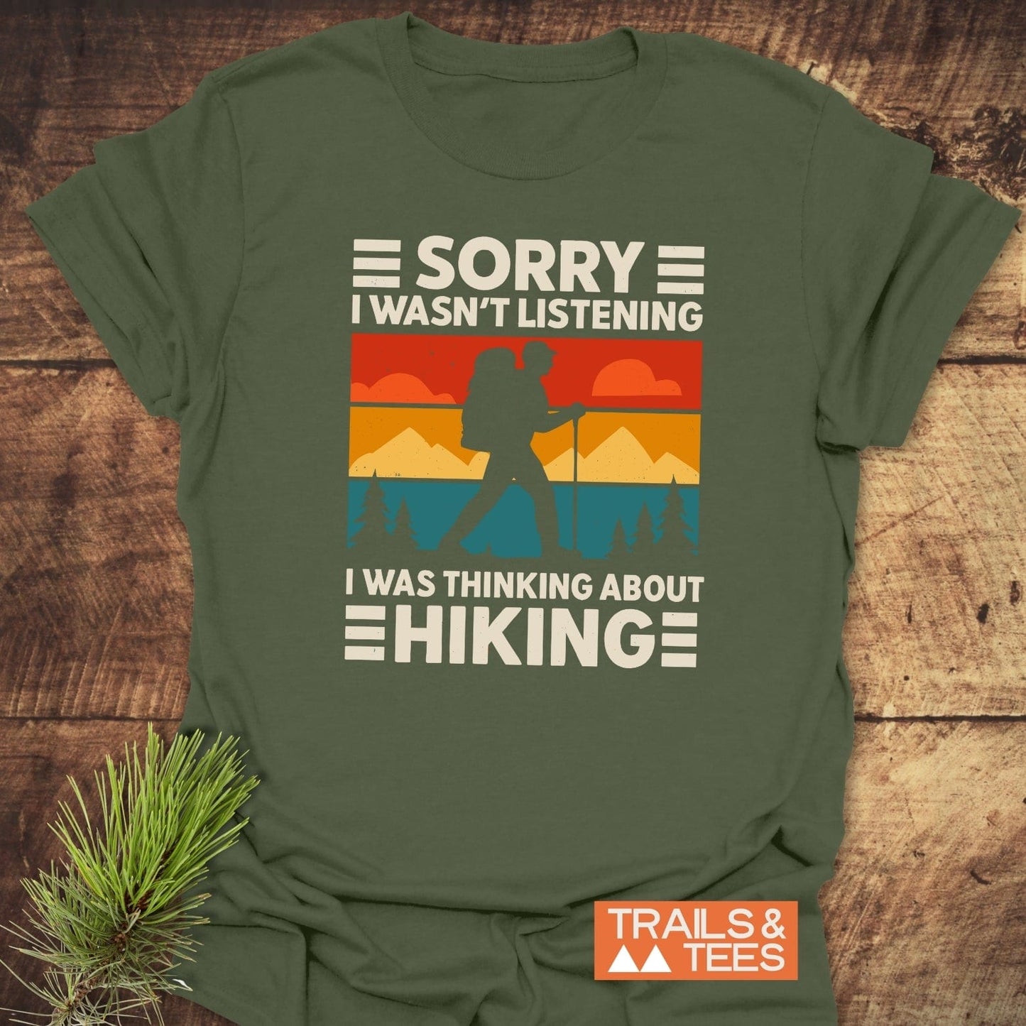Thinking About Hiking T-Shirt featuring a retro backpacker graphic, sunset, and pine trees, laid on wood with pine sprig; Trails & Tees logo visible.