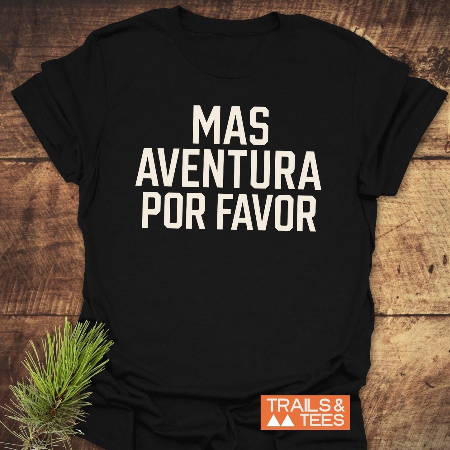 Mas Aventura T-Shirt on wood, featuring bold white text and small orange logo, with pine needles beside, highlighting outdoor theme and adventure spirit.