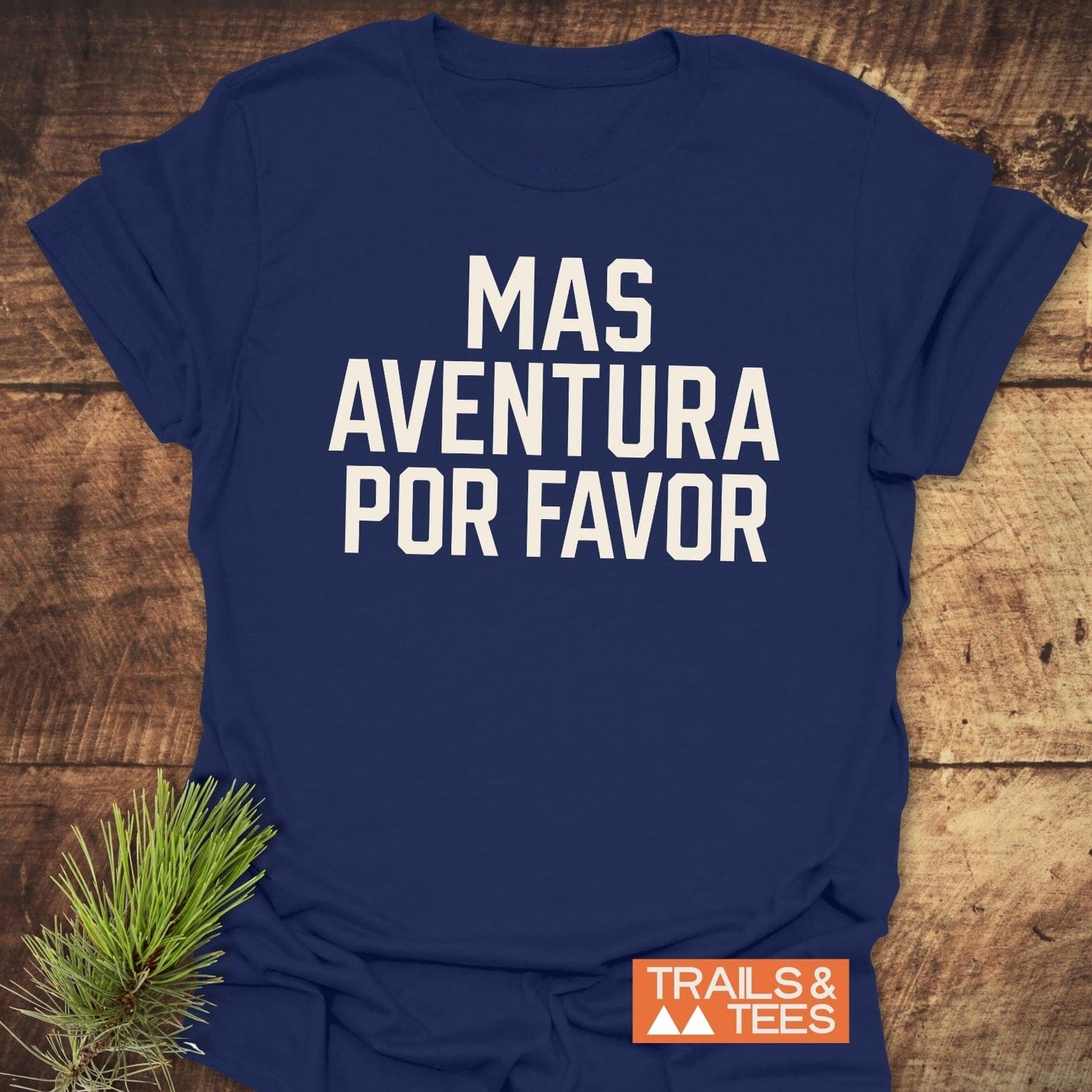 Mas Aventura T-Shirt with “MAS AVENTURA POR FAVOR” in white lettering, styled with a pine sprig and featuring an orange “TRAILS & TEES” logo.