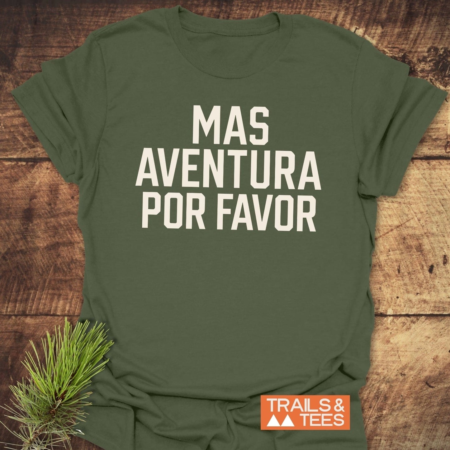 Mas Aventura T-Shirt on rustic wood, bold lettering, orange Trails & Tees badge, pine needles beside it; lightweight cotton, adventure-ready design.