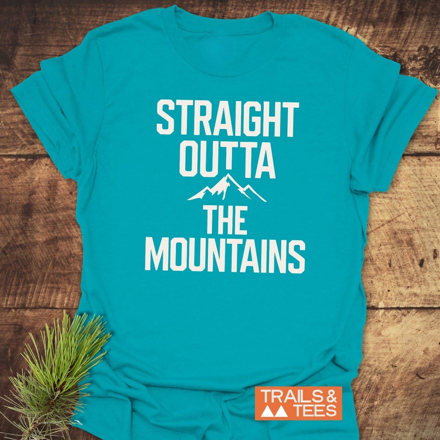 Straight Outta The Mountains T-Shirt with bold white text, orange logo, on soft cotton, laid flat with pine sprig accent beside.