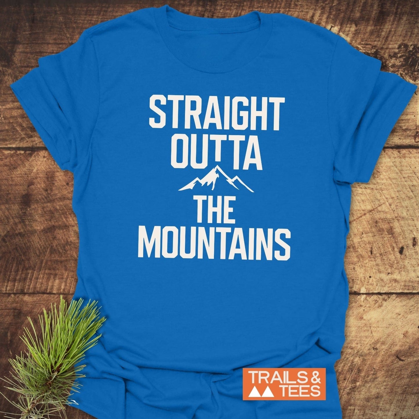 Straight Outta The Mountains T-Shirt in soft cotton with slogan and mountain graphic, featuring Trails & Tees logo, styled with pine sprig on rustic wood.