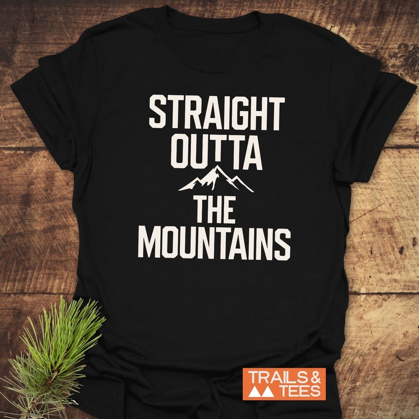 Straight Outta The Mountains T-Shirt on rustic wood, featuring bold white text with mountain outline. Includes pine sprig accent, emphasizing outdoor theme.
