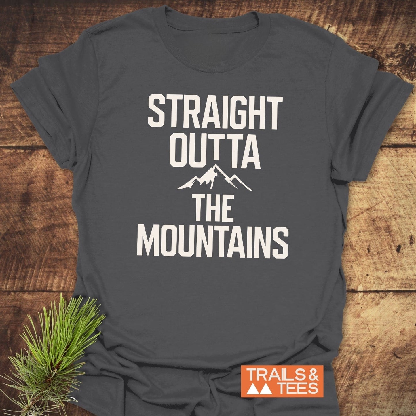 Straight Outta The Mountains T-Shirt on wood, with white block print and mountain silhouette; includes Trails & Tees logo and pine sprig accent.