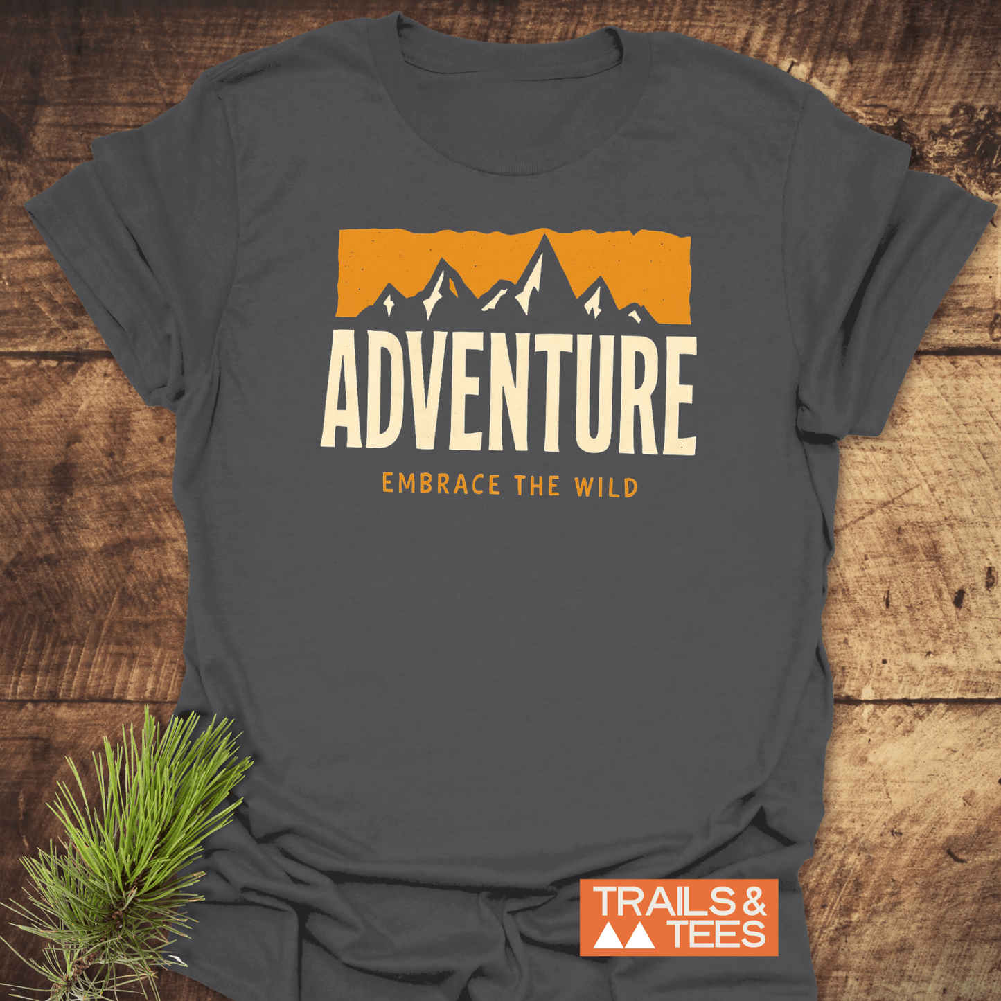 Adventure T-Shirt with mountain graphic, ADVENTURE text, and EMBRACE THE WILD slogan, featuring durable print on soft, pre-shrunk cotton, displayed on a wooden surface.