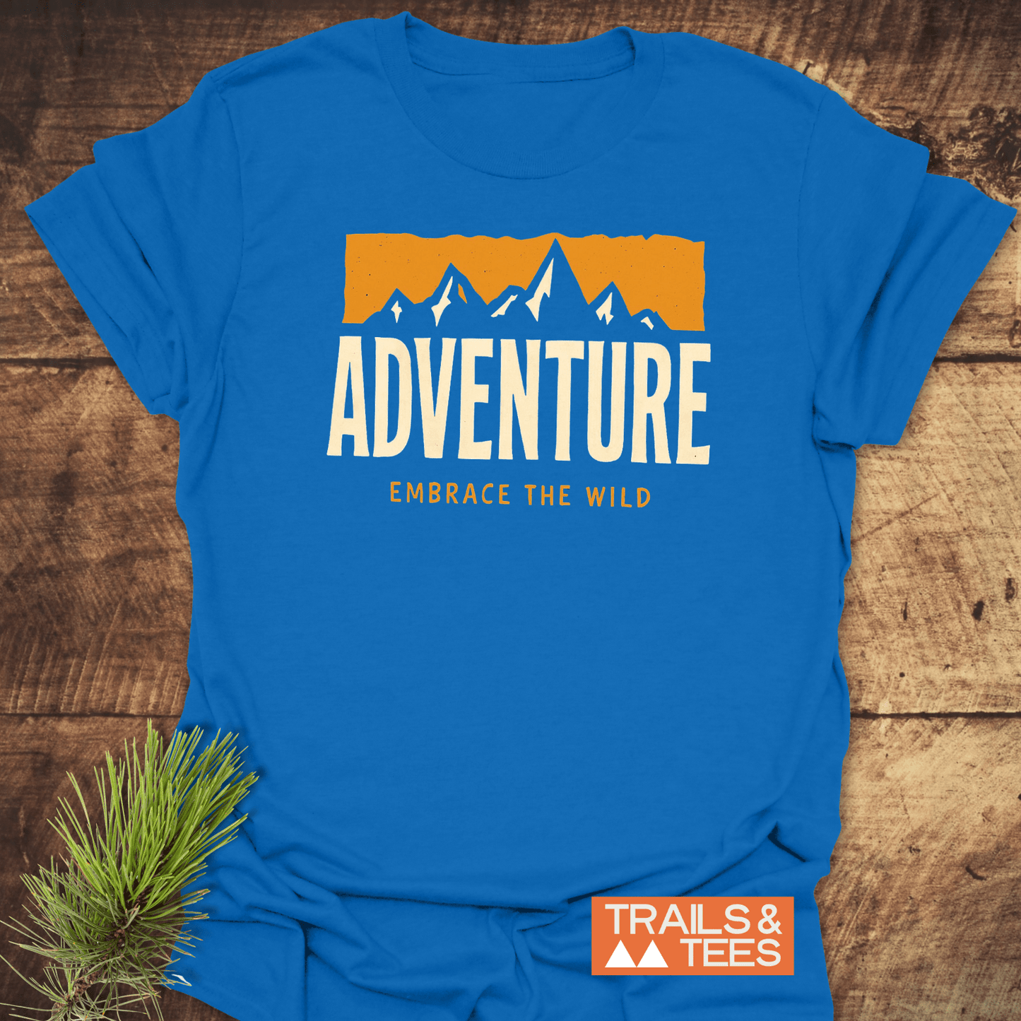 Adventure T-Shirt featuring a mountain-range graphic with ADVENTURE and EMBRACE THE WILD text, laid on wood, showcasing outdoor-inspired design.