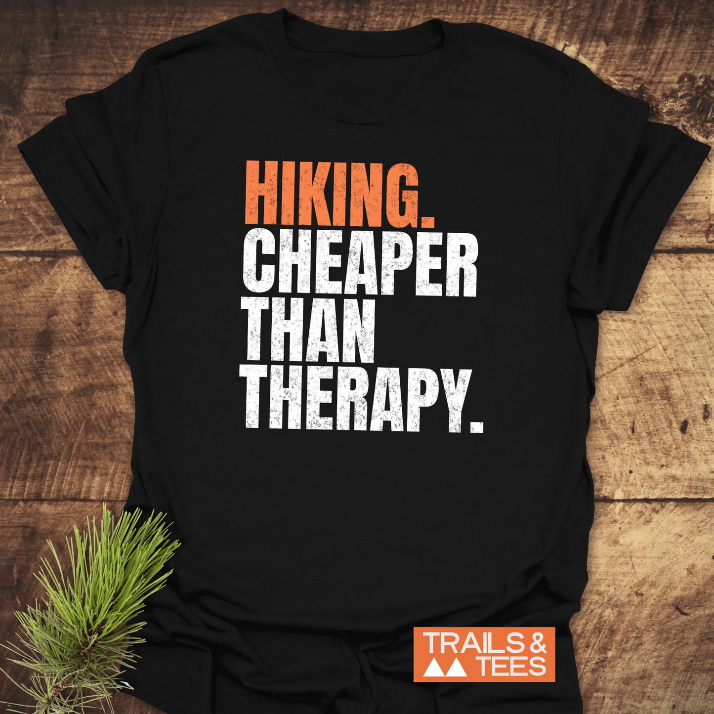 Black Hiking. Cheaper Than Therapy T-shirt on wood, with pine sprig. Features distressed lettering, durable print, and TRAILS & TEES logo.