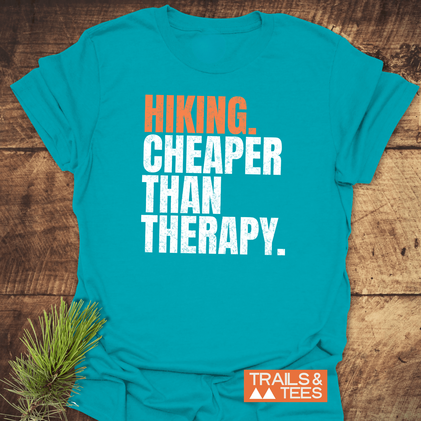 Teal-blue Hiking. Cheaper Than Therapy T-shirt on wood, features orange Trails & Tees badge with mountains, beside a pine sprig.