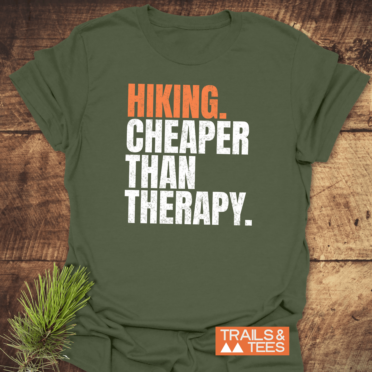 Olive-green Hiking. Cheaper Than Therapy T-shirt on wooden surface, featuring distressed letters and TRAILS & TEES logo, crafted from soft, durable cotton.