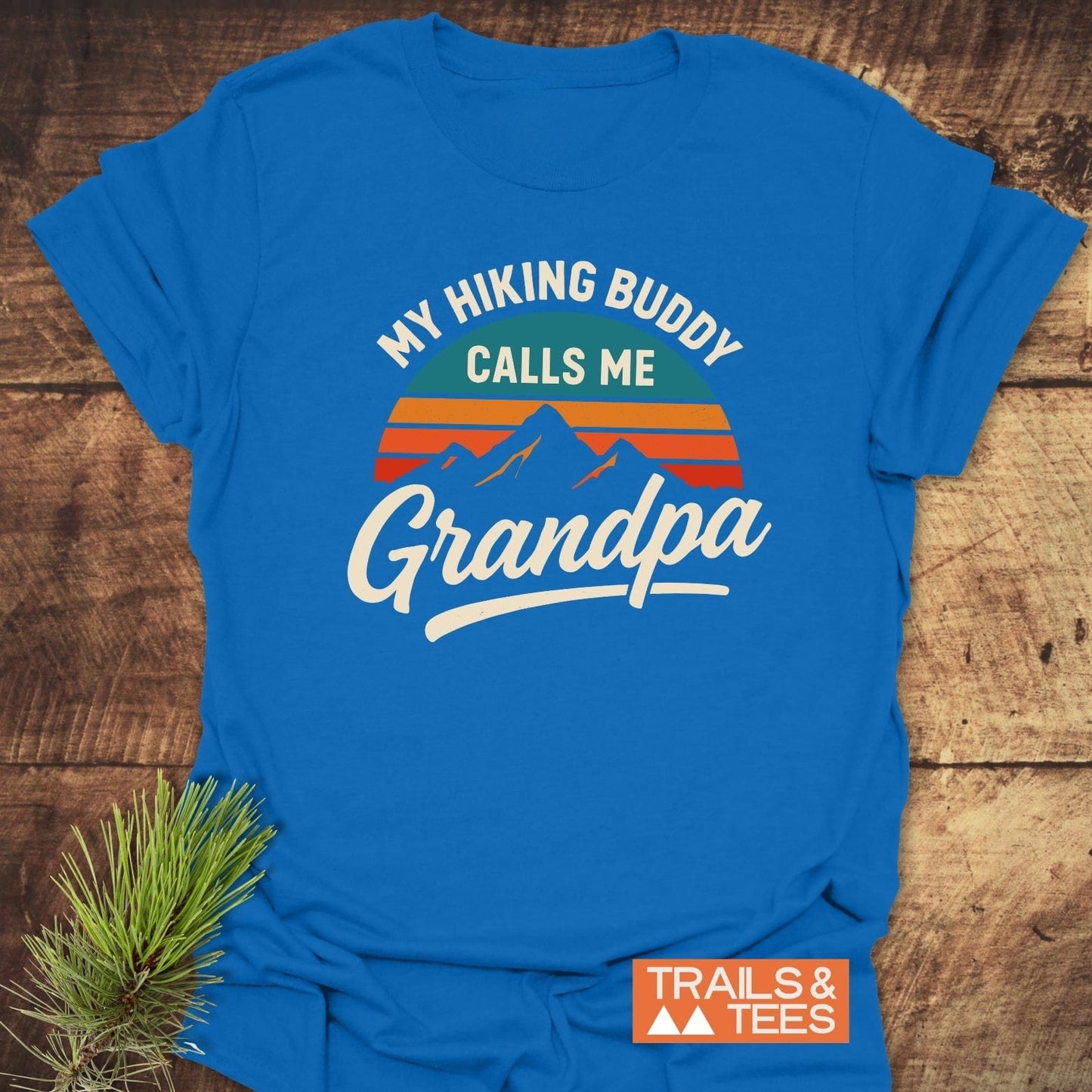 Royal-blue Grandpa Hiking Buddy T-Shirt with stylized mountain and sun graphic, laid flat on wood, highlighting outdoor adventure theme and durable, lightweight cotton fabric.