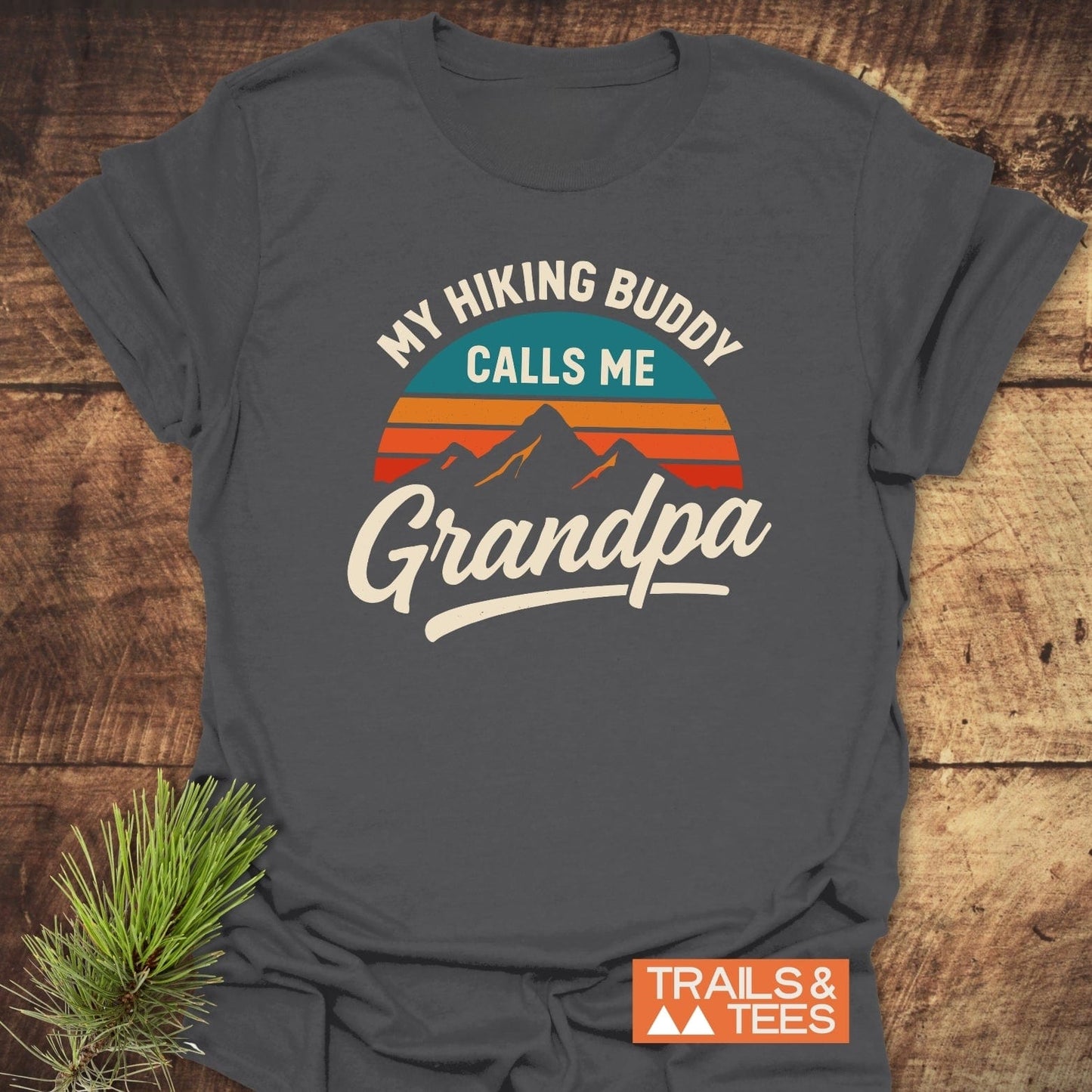 Grandpa Hiking Buddy T-Shirt with mountain graphic and slogan on dark-gray fabric, laid on wood. Soft, durable cotton ideal for outdoor enthusiasts.