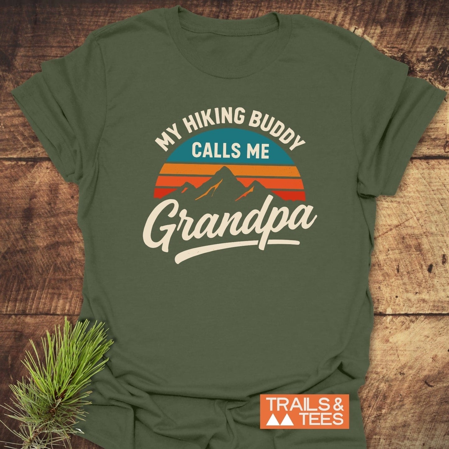 Grandpa Hiking Buddy T-Shirt on wood, featuring a mountain-sunset graphic. Made from soft, pre-shrunk cotton with durable prints, ideal for outdoor enthusiasts.
