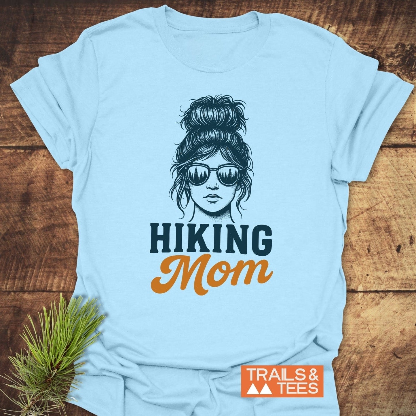 Messy Bun Hiking Mom T-Shirt featuring a woman with sunglasses, reflecting a treeline, and the words “HIKING Mom.” Includes durable prints and taped shoulders.