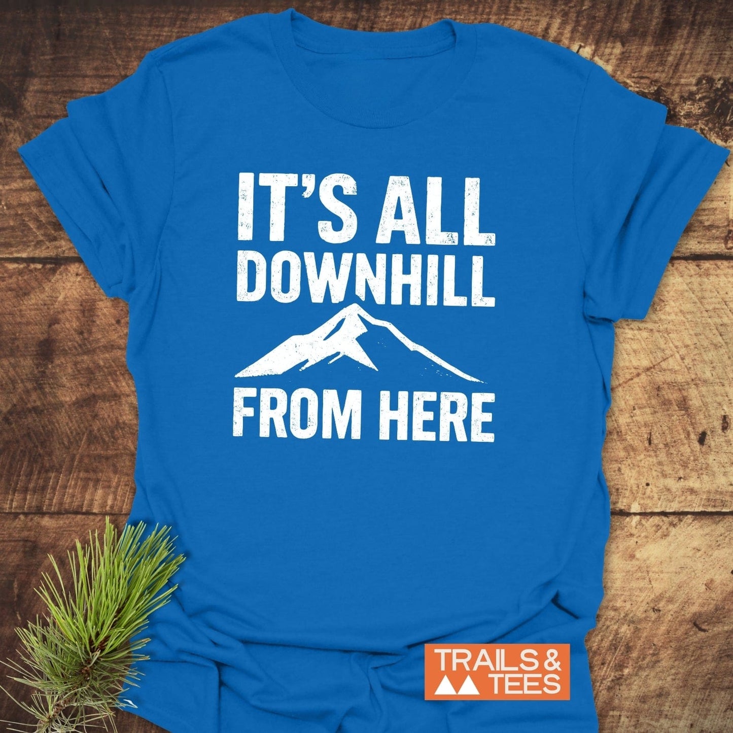 All Downhill T-Shirt on wood, featuring bold white text and mountain silhouette. Includes orange TRAILS & TEES logo, emphasizing adventure-ready design and comfort.