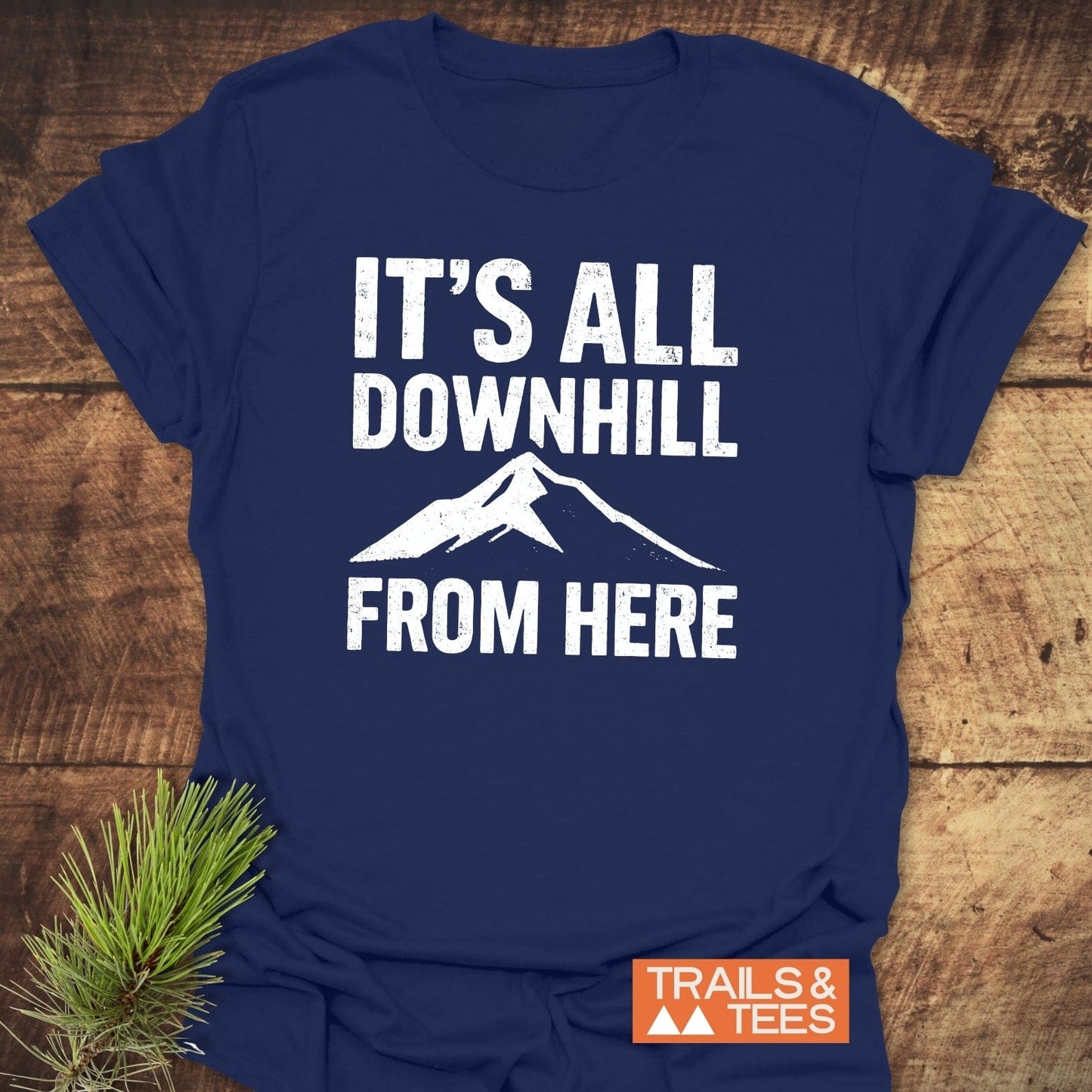 All Downhill T-Shirt on wooden surface, featuring a mountain silhouette and slogan. Includes Trails & Tees logo. Perfect for outdoor adventures.