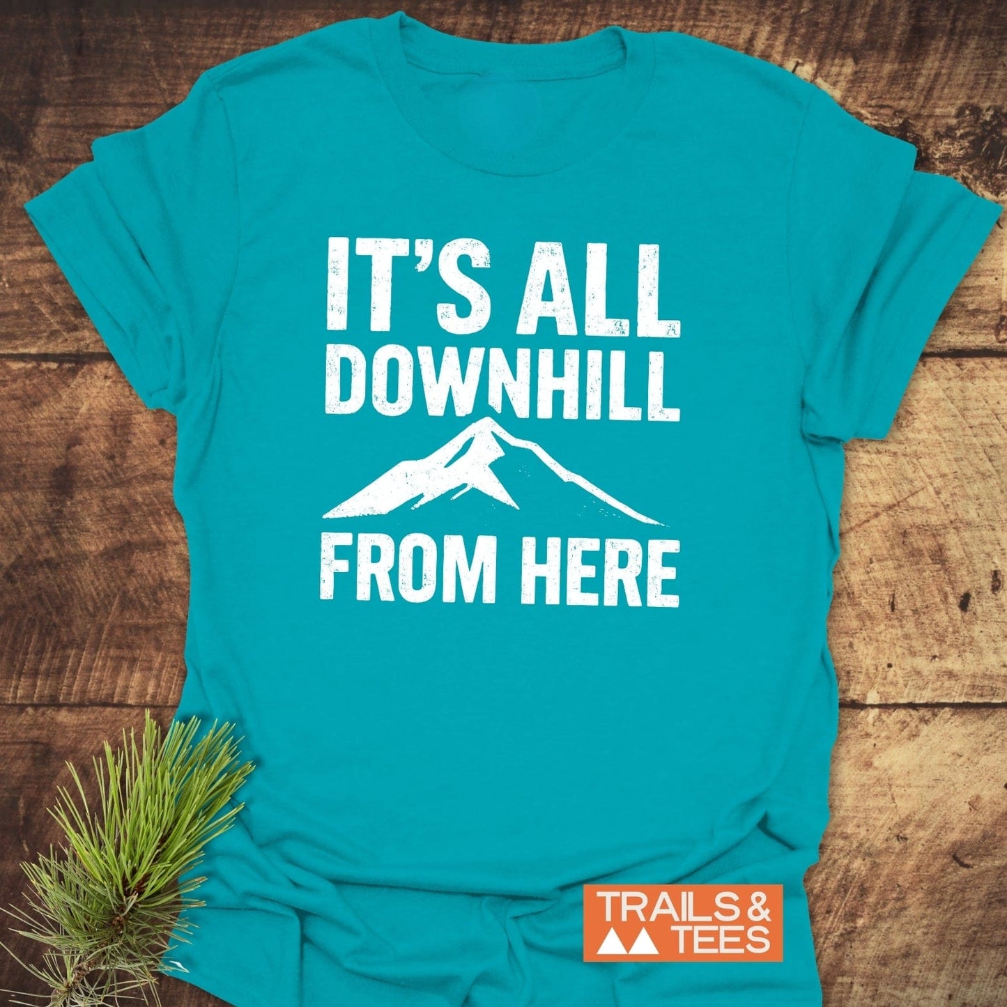 All Downhill T-Shirt featuring a mountain-themed design, large white text, and orange label, laid on wood surface with pine needles.
