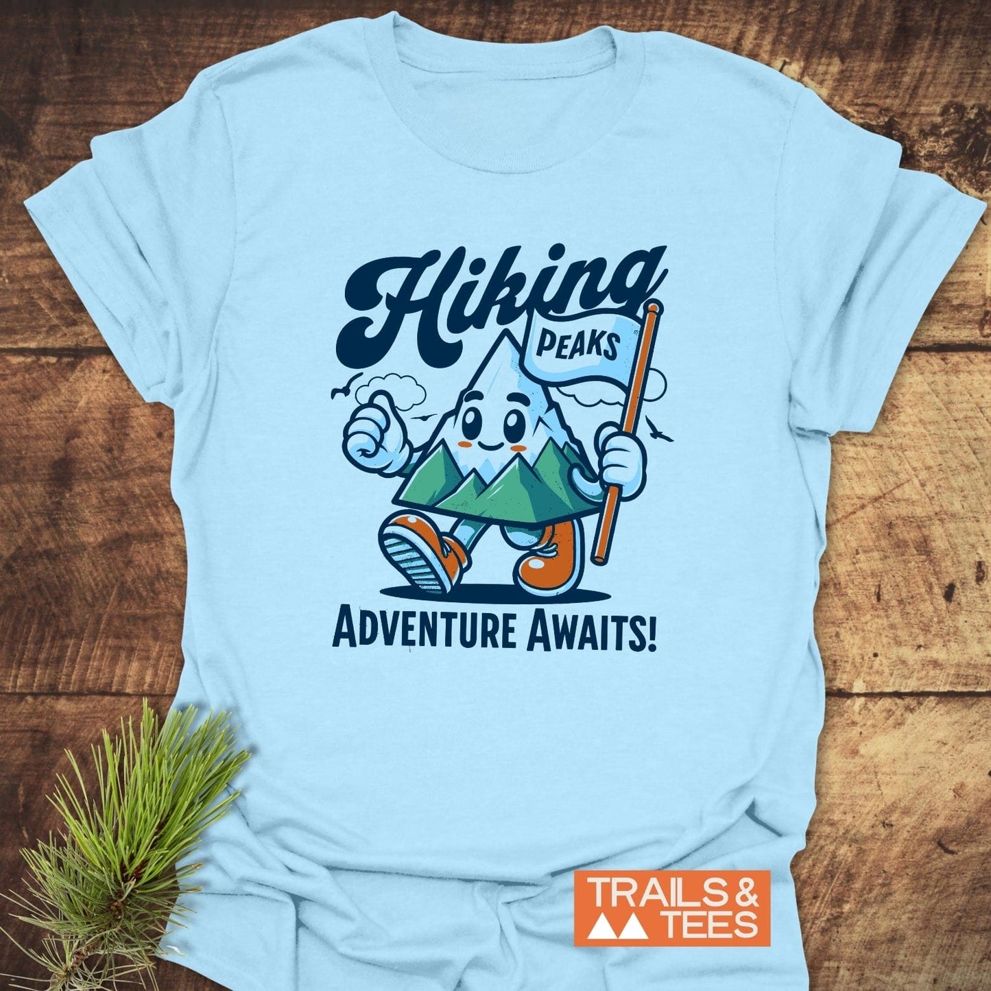Light-blue Hiking Time Funny T-Shirt with cartoon mountain graphic, Hiking Peaks text, and Adventure Awaits! slogan, featuring Trails & Tees logo on a wooden surface.