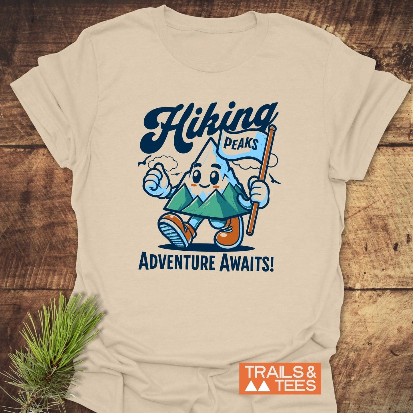 Hiking Time Funny T-Shirt: Cream tee with cartoon mountain character, Hiking script, Adventure Awaits! slogan, TRAILS & TEES logo, laid on wood with pine sprig.
