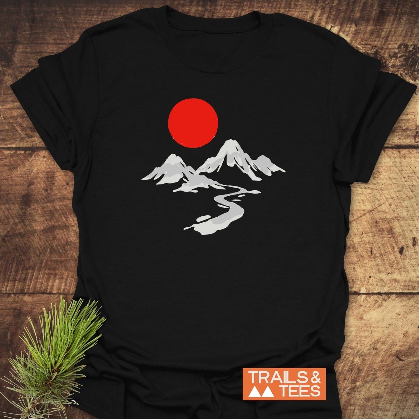 Crimson Sun T-Shirt featuring a minimalist outdoor scene with mountains, a river, and a red sun; styled on wood with pine sprig and Trails & Tees logo.