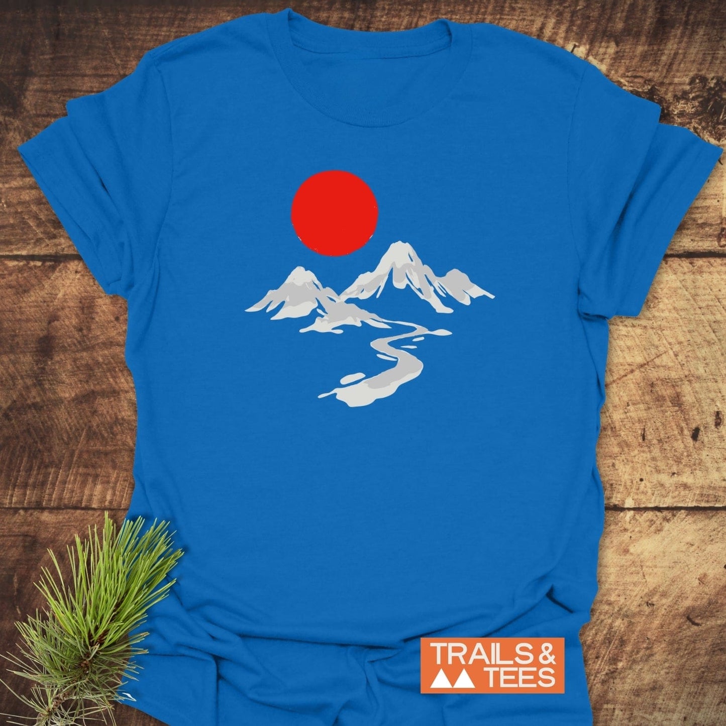 Crimson Sun T-Shirt featuring a minimalist mountain scene with snow-topped peaks and red sun. Includes TRAILS & TEES logo; styled on rustic wood with pine sprig.