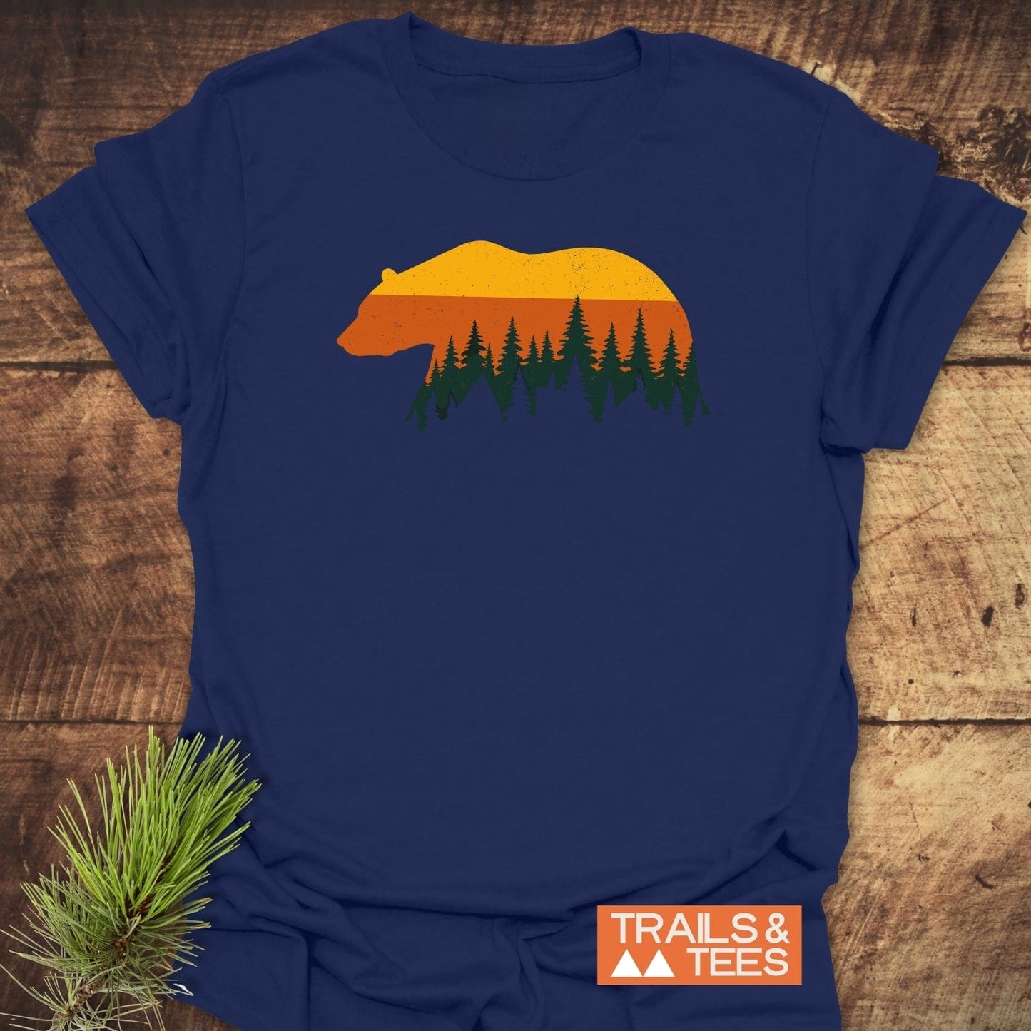 Vintage Grizzly Sunset T-Shirt featuring a bear silhouette with a retro sunset and fir trees graphic, displayed on a wooden surface with TRAILS & TEES logo.