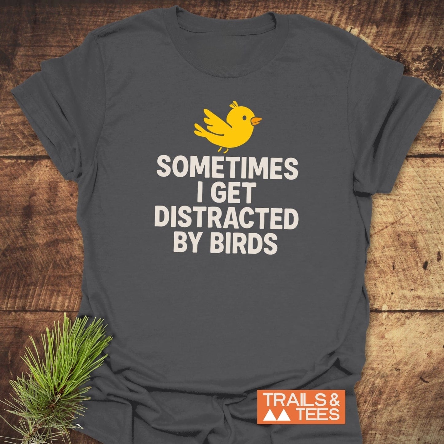 Bird Distraction Funny T-Shirt with a yellow cartoon bird and the phrase SOMETIMES I GET DISTRACTED BY BIRDS, laid on a wooden surface with pine sprig styling.