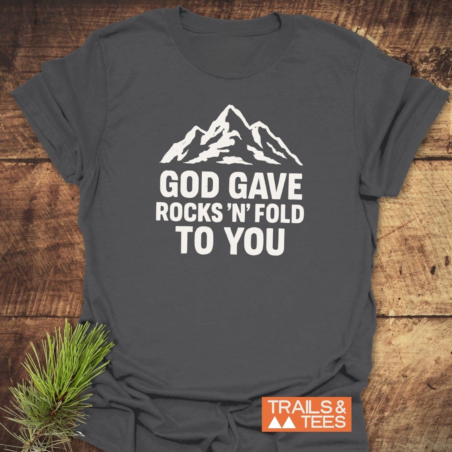Charcoal-gray T-shirt with a white mountain silhouette and God Gave Rocks ’N’ Fold To You, features TRAILS & TEES logo and pine branch accent.