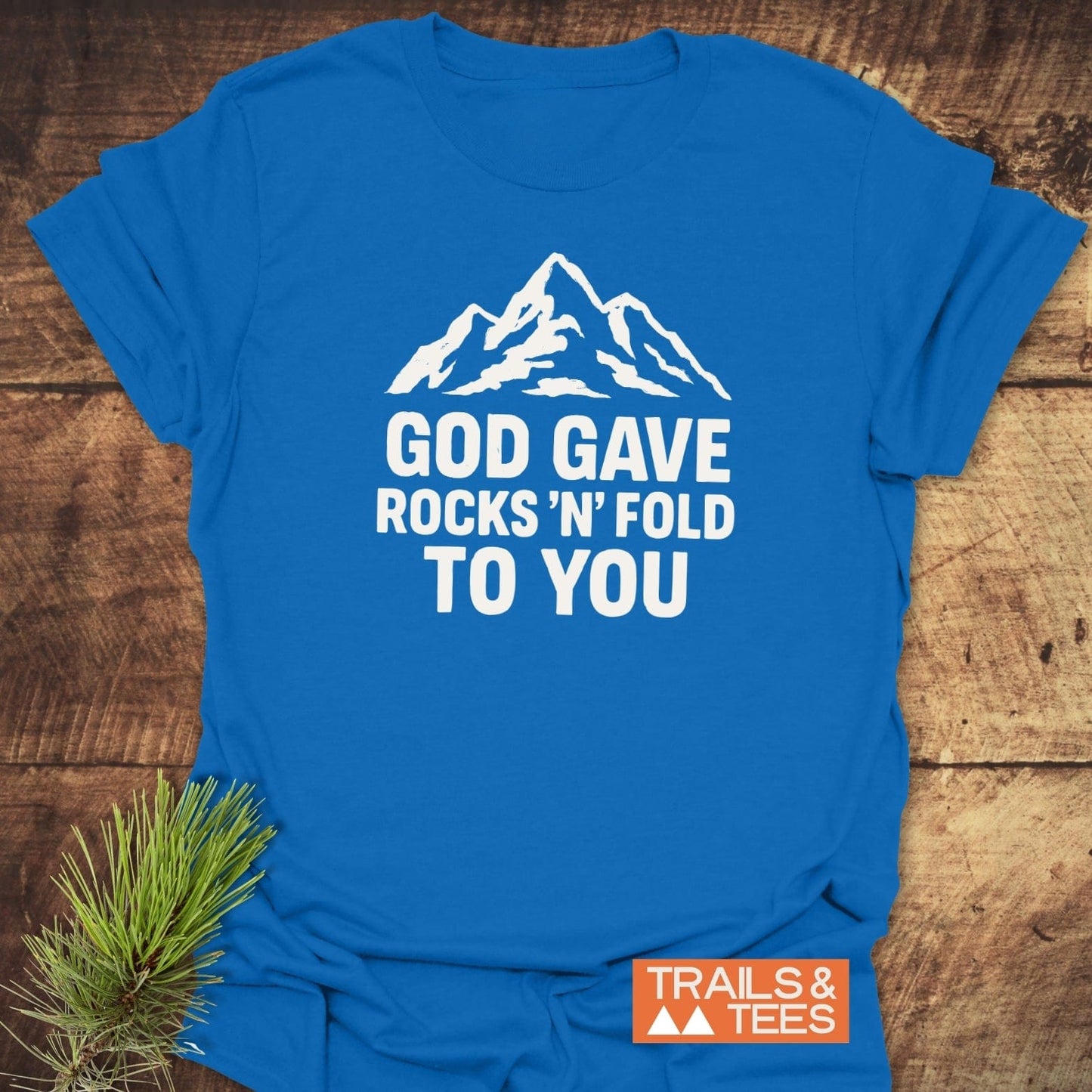 Bright blue God Gave... Funny Geology T-Shirt with mountain graphic and slogan, featuring TRAILS & TEES logo, on wooden surface with pine needles.