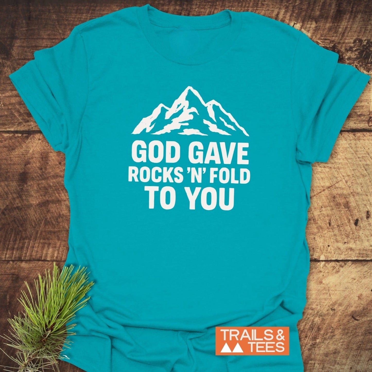 Turquoise t-shirt with mountain silhouette and “GOD GAVE ROCKS ’N’ FOLD TO YOU” text. Features “TRAILS & TEES” logo, highlighting adventure-ready design.