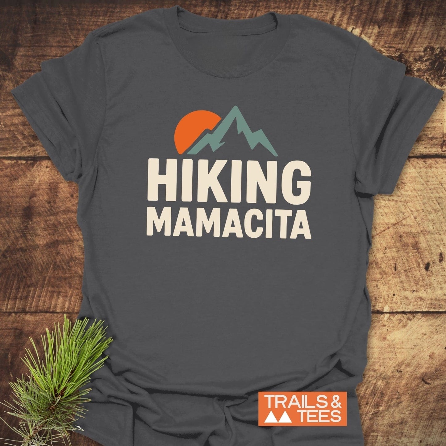 Charcoal-gray Hiking Mamacita T-shirt with stylized mountains and sun graphic, crafted from soft cotton, laid flat on a wooden surface with pine sprig.