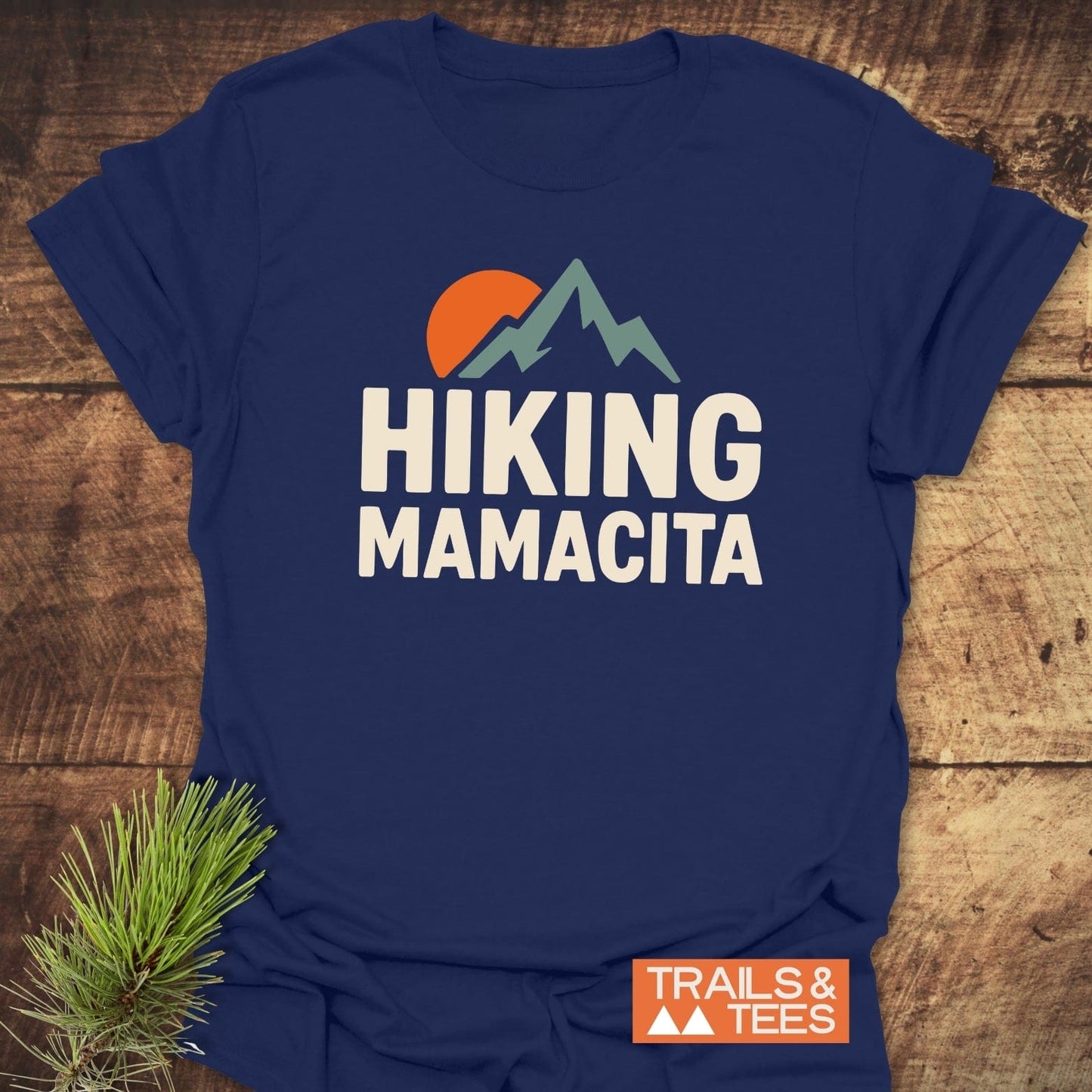 Navy-blue Hiking Mamacita T-shirt with mountain graphic and sun, featuring orange TRAILS & TEES logo. Soft, durable, pre-shrunk cotton, ideal for outdoor adventures.