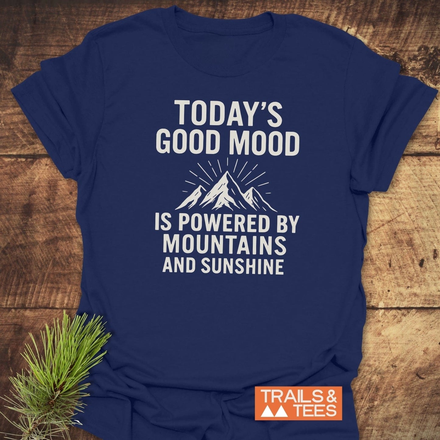 Powered By Mountains T-Shirt on wood, featuring mountain graphic and slogan. Includes Trails & Tees logo. Made of soft cotton for comfort and durability.