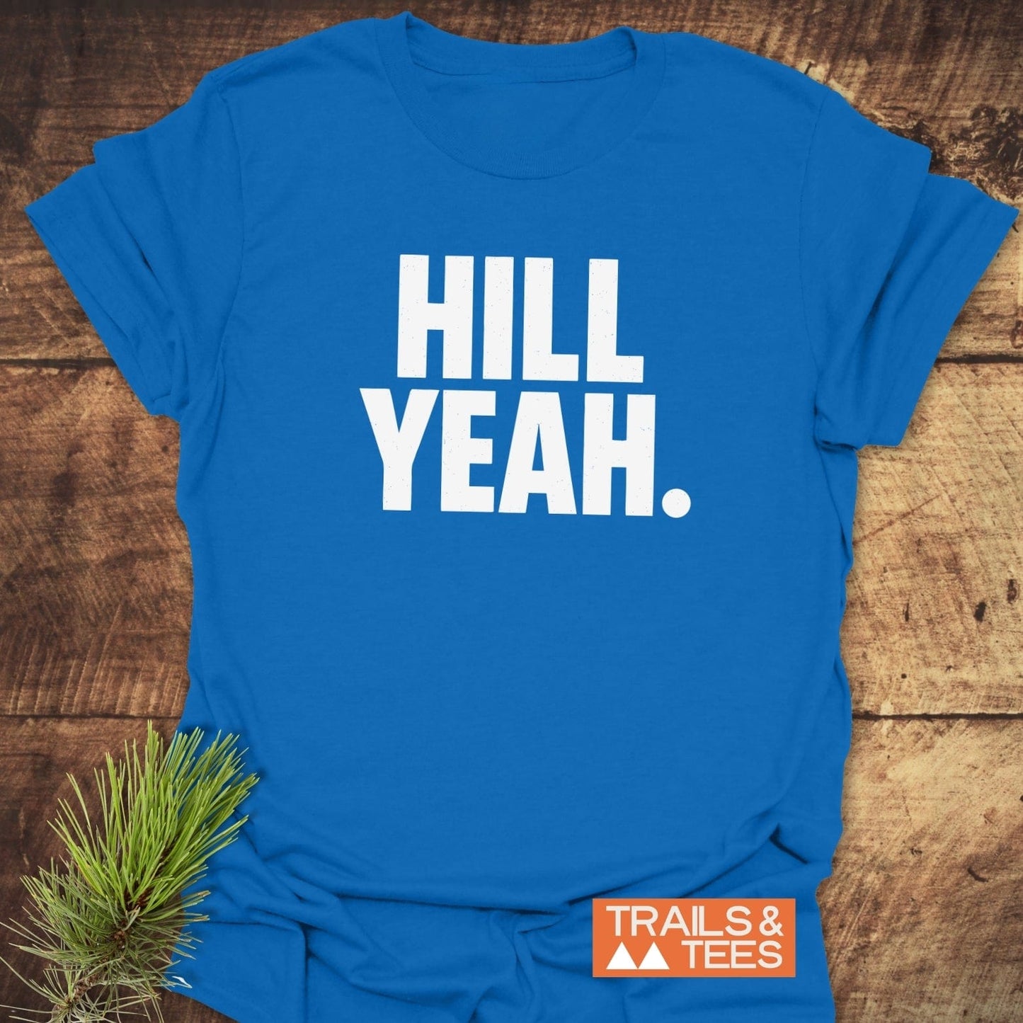 Hill Yeah T-Shirt laid flat; bright blue with bold white lettering, orange logo tag, and pine sprig. Soft, durable cotton ideal for outdoor adventures.