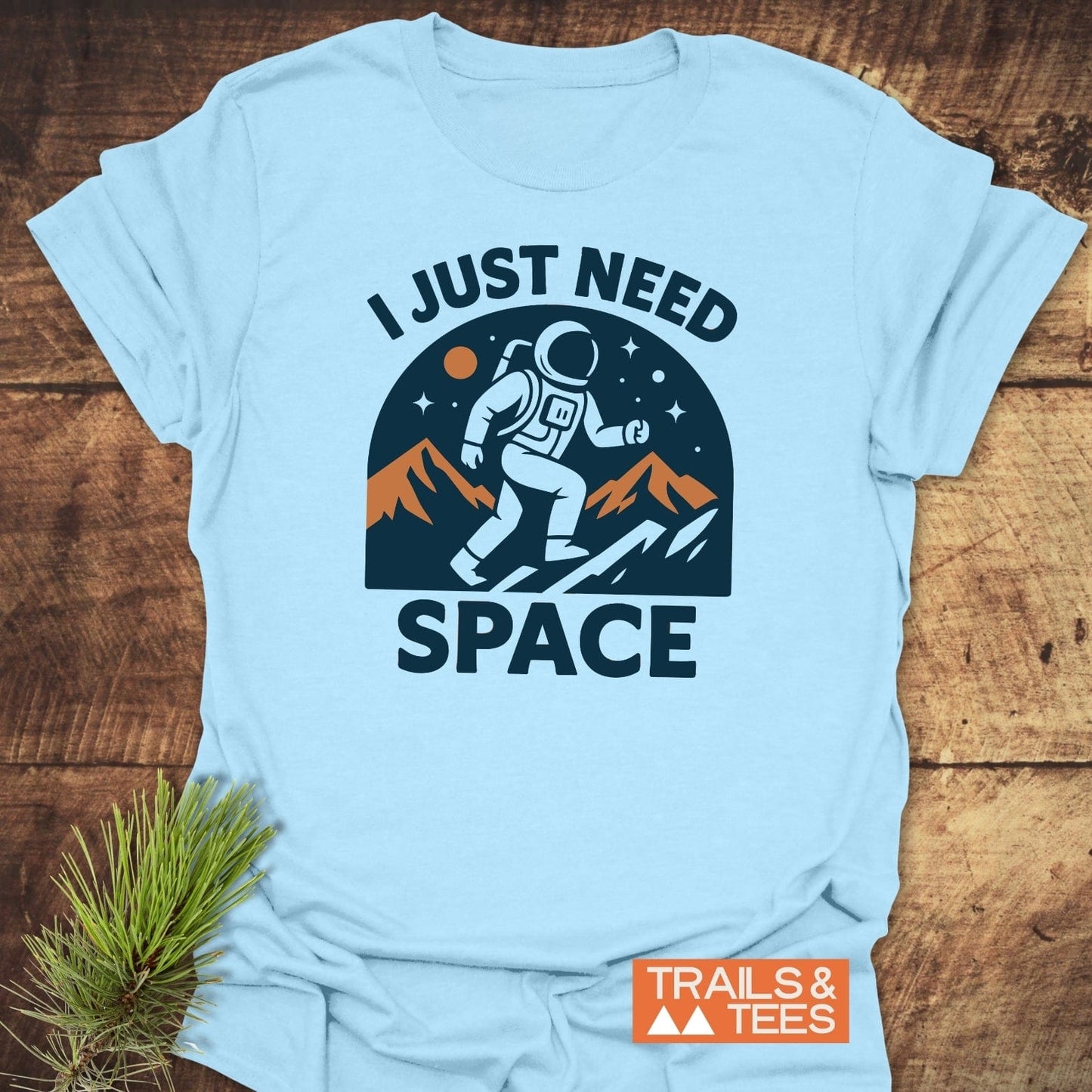 Pale-blue I Just Need Space T-shirt with astronaut graphic, rust/orange mountains, and starry sky. Ideal for outdoor enthusiasts, crafted from soft, durable cotton.