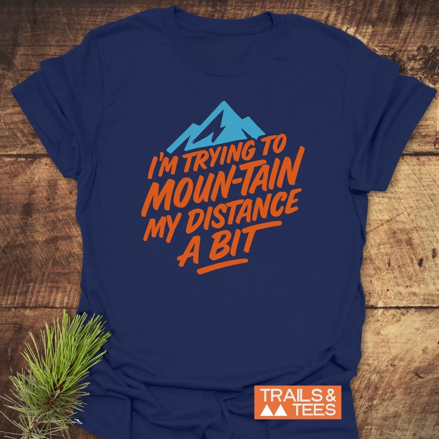 Navy-blue Moun-Tain Distance Funny T-Shirt with mountain graphic and orange text, laid on wood. Features comfortable, durable cotton, ideal for outdoor enthusiasts.