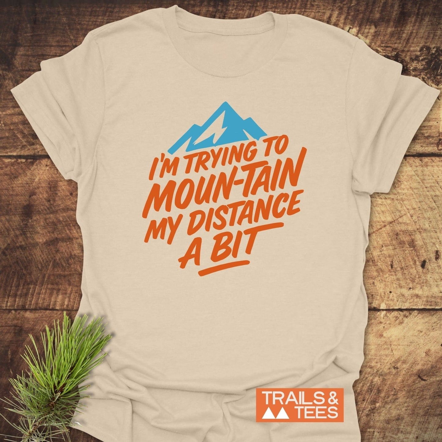 Moun-Tain Distance Funny T-Shirt on wood, featuring a blue mountain and bold orange text. Soft cotton, durable prints, designed for outdoor enthusiasts.