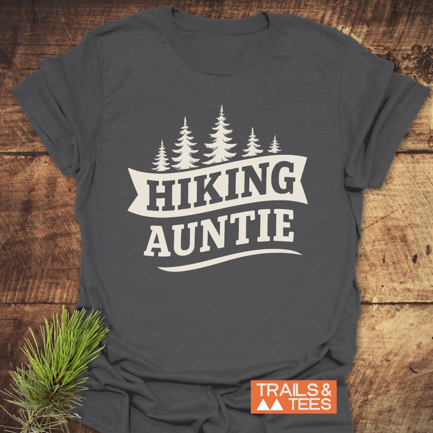 Hiking Auntie T-Shirt with pine tree graphic and “TRAILS & TEES” label, set on wood with pine branch, highlights durability and outdoor adventure appeal.