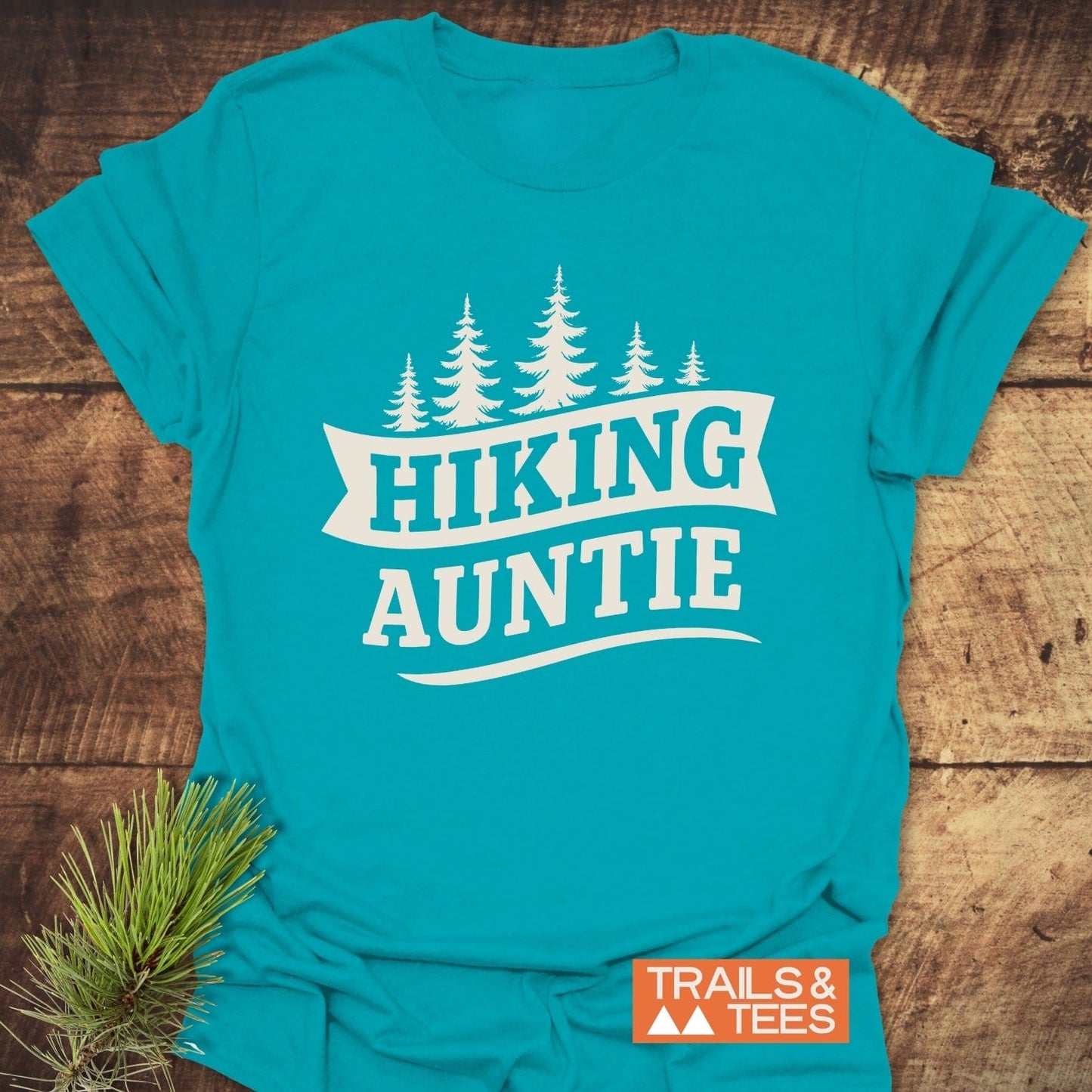 Teal Hiking Auntie T-shirt with white pine tree graphic, crafted from soft cotton, laid on wood surface with pine branch prop.