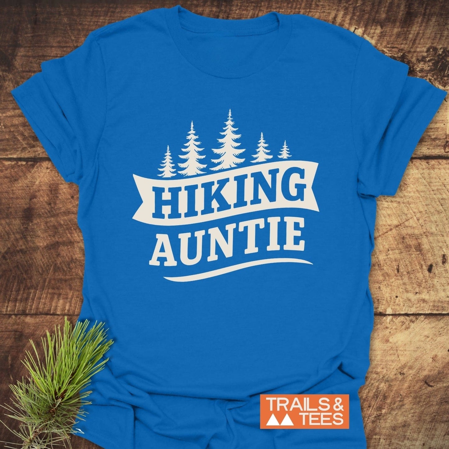 Hiking Auntie T-Shirt on wood, featuring pine silhouettes and logo. Soft, lightweight cotton for adventure lovers; durable print, pre-shrunk fit, eco-friendly production.
