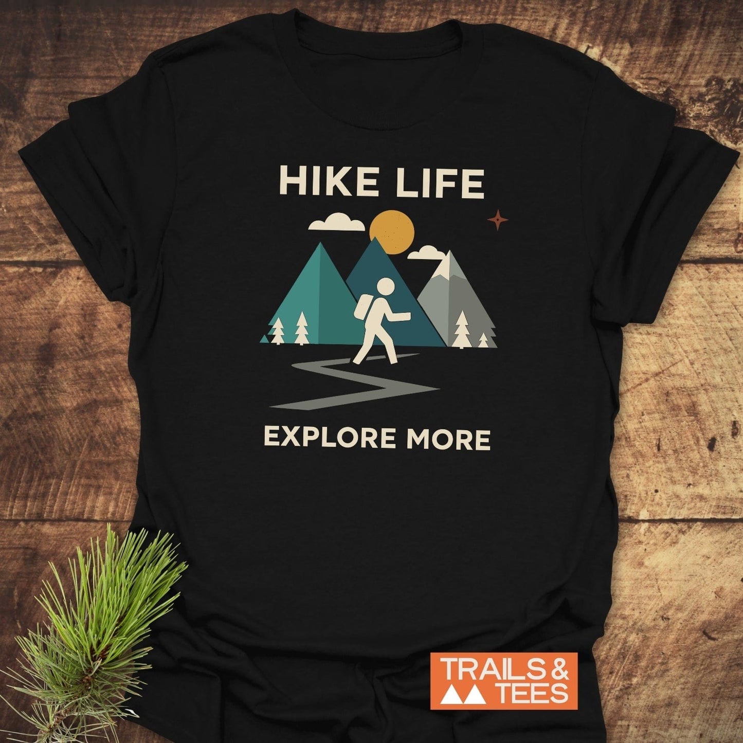 Hike Life Graphic T-Shirt with mountain scene, hiker, and EXPLORE MORE slogan, styled with a pine sprig on a wooden surface.