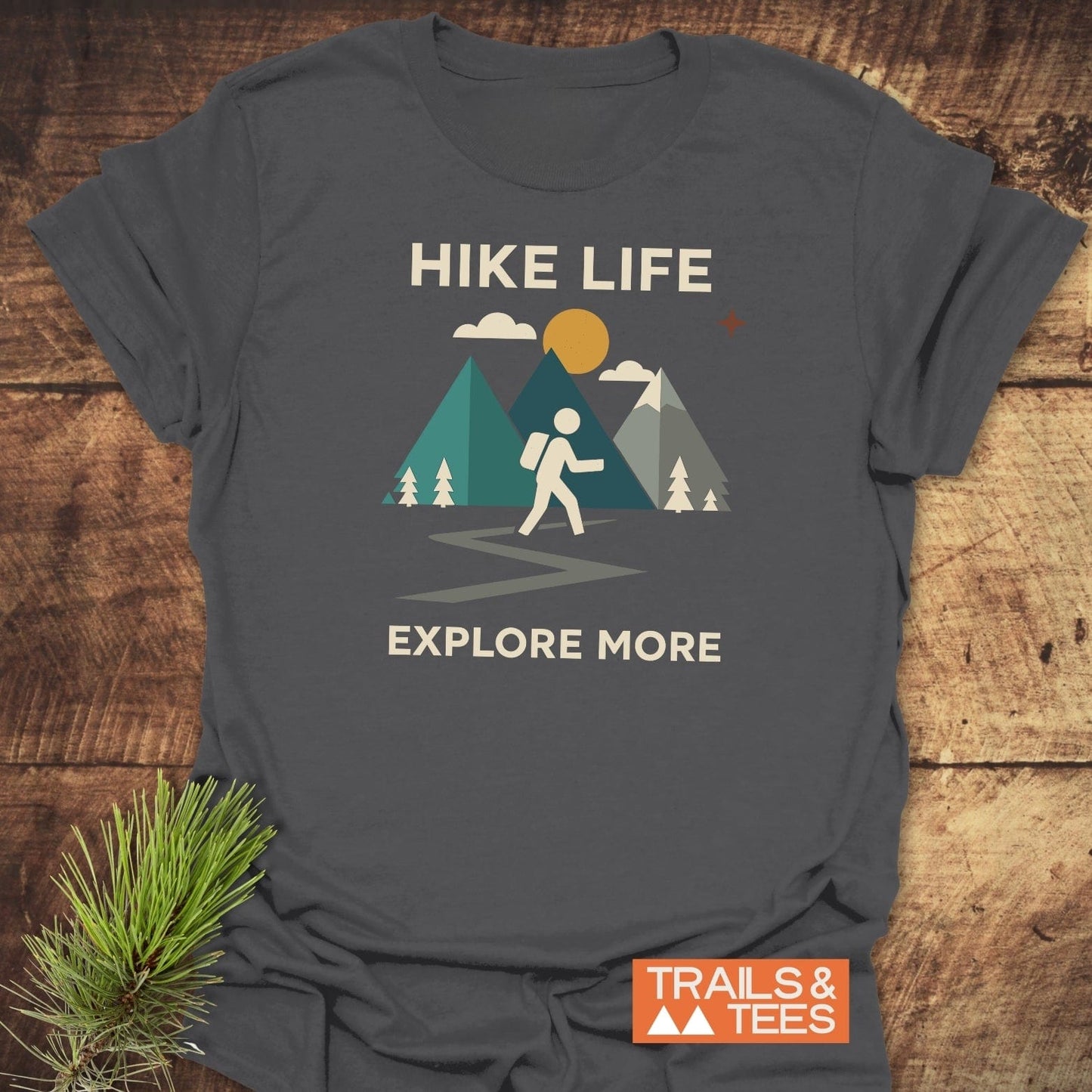 Hike Life Graphic T-Shirt featuring a stick-figure hiker, mountains, and sun graphic, with EXPLORE MORE text and TRAILS & TEES logo on a dark-gray tee.