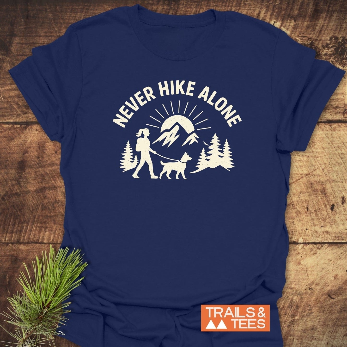 Never Hike Alone Dog Mom T-Shirt showing a hiker with a dog, pine trees, mountains, and sun-rays, featuring the TRAILS & TEES logo and a rustic pine branch.