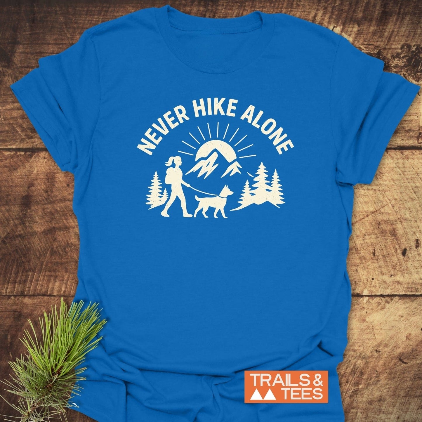 Never Hike Alone Dog Mom T-Shirt features a hiker with dog, pine trees, and mountains. Made from soft, durable cotton with DTF prints, ensuring lasting quality.