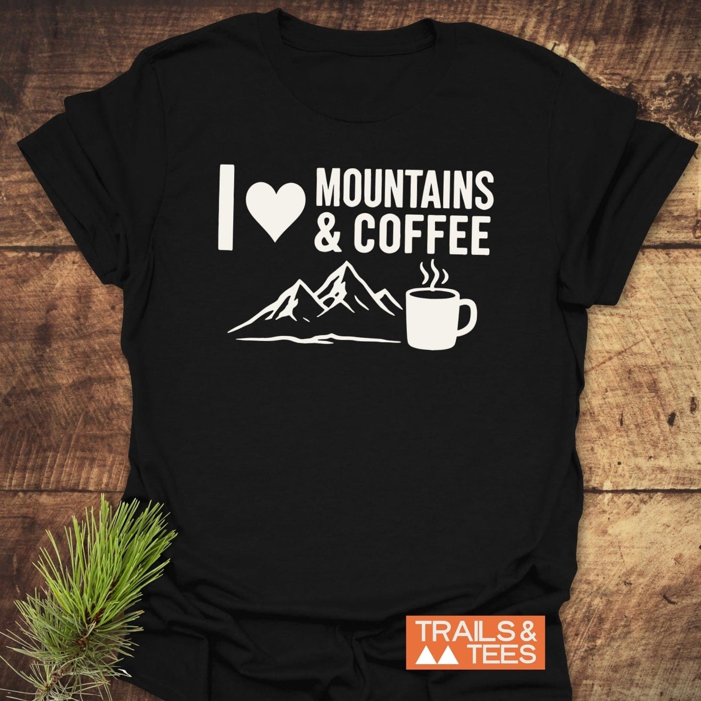 I love Mountains And Coffee T-Shirt with mountain and coffee mug graphics, featuring durable prints, laid on a wooden surface with a pine sprig.
