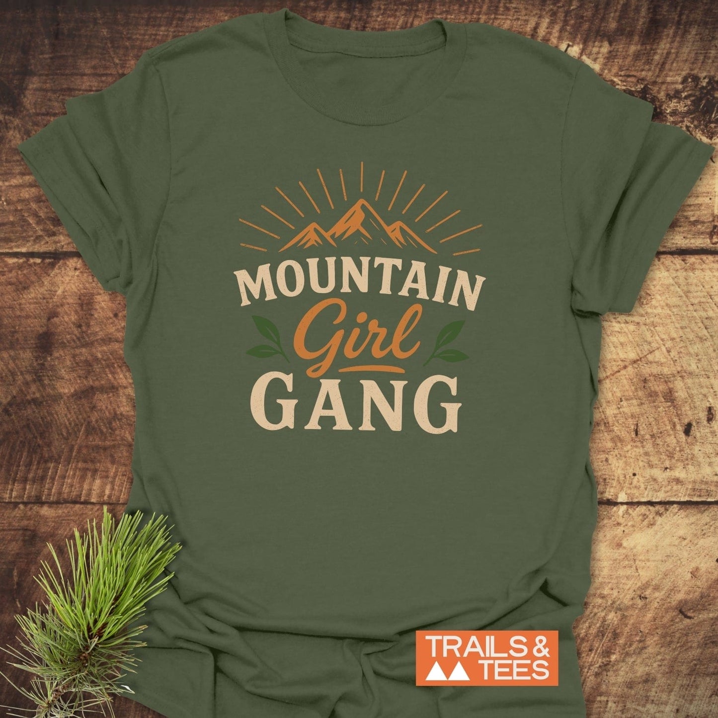 Girl Gang Hiking T-Shirt with mountain graphic and MOUNTAIN Girl GANG text, featuring TRAILS & TEES label; accented by evergreen sprig on wood background.