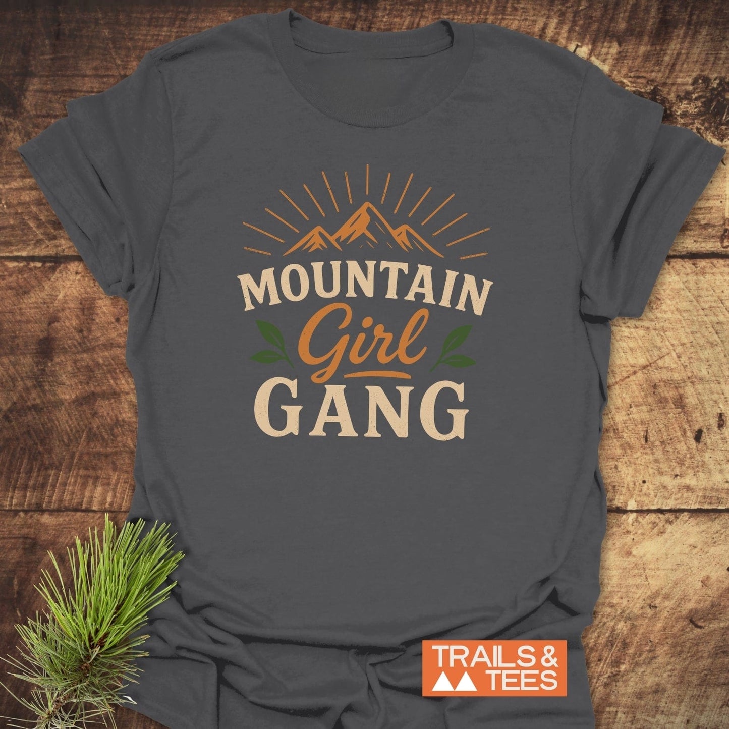 Girl Gang Hiking T-Shirt featuring sun-lit mountains graphic and “MOUNTAIN Girl GANG” text, laid on wood with a pine sprig and “TRAILS & TEES” logo.