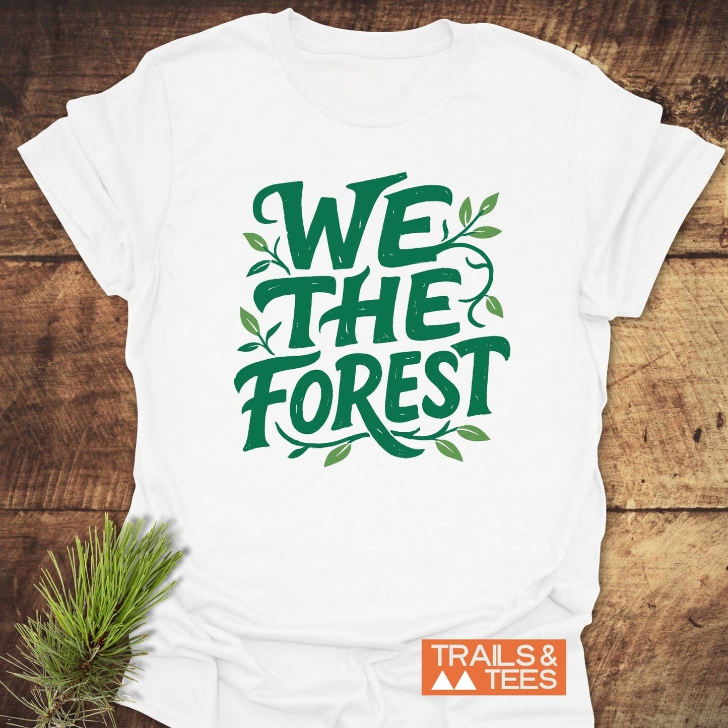 We The Forest T-Shirt with green slogan and leaf details, laid on wood. Features durable print, soft cotton, pre-shrunk fit, and eco-friendly production.