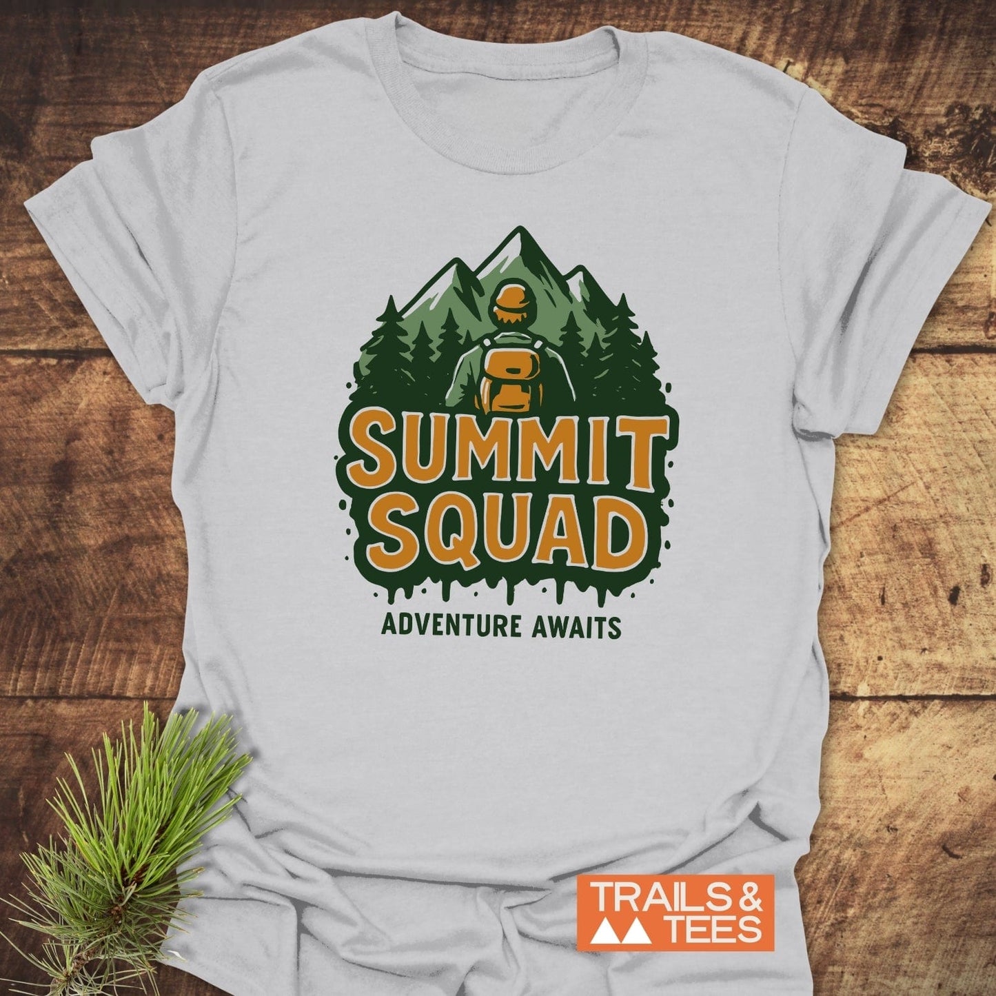Summit Squad Hiking T-Shirt featuring a hiker graphic and adventure slogan. Light cotton, DTF print, laid flat with pine sprig. Ideal for outdoor enthusiasts.