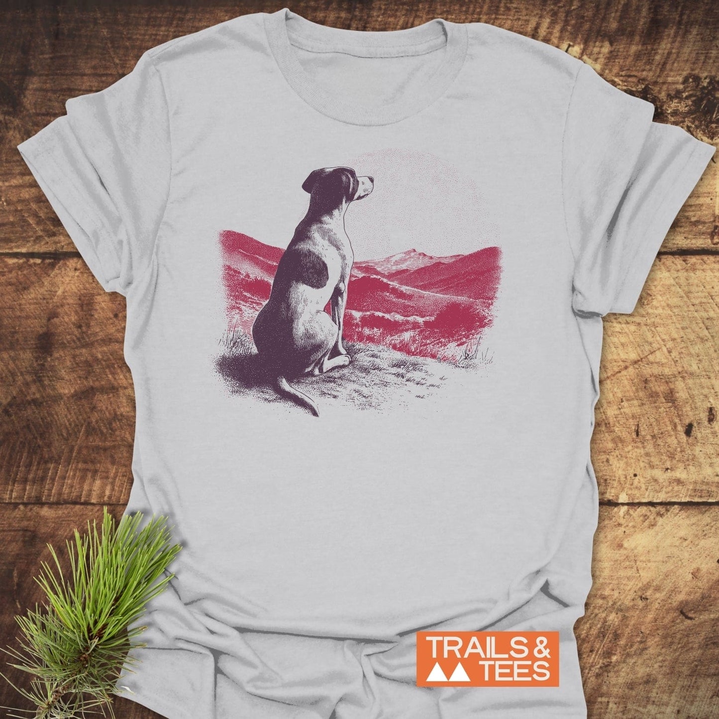 Mountains and Dog T-Shirt featuring a dog illustration over a mountain landscape, with TRAILS & TEES logo. Laid on wood surface with pine sprig.