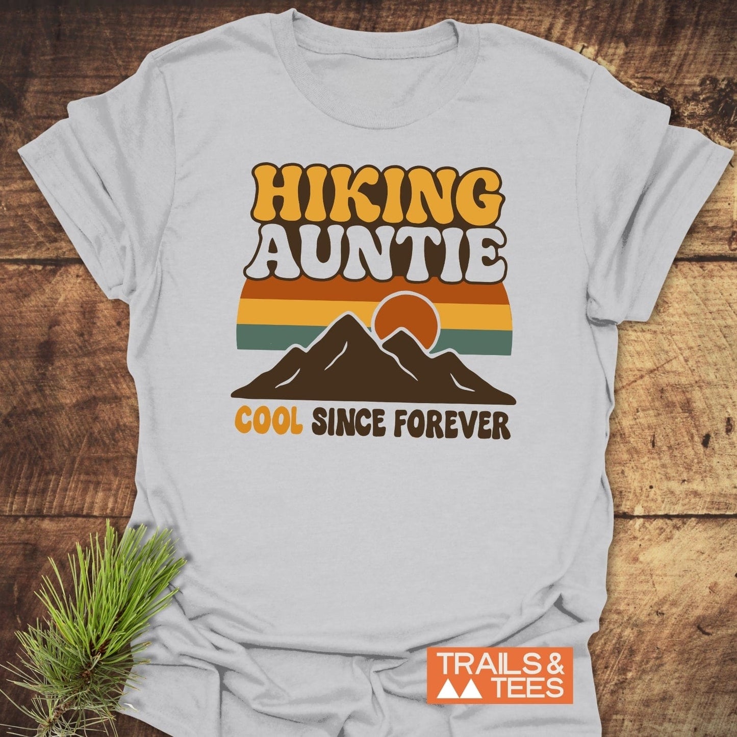 Cool Hiking Auntie T-Shirt with retro mountain graphic and text, laid on wood. Features soft, ring-spun cotton, pre-shrunk, durable print.